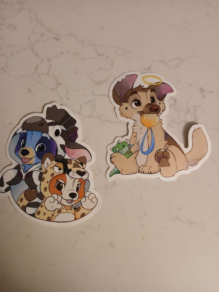 A pair of stickers made by Madshyarts. One is of a puppy-sized Charlie, from "All Dogs go to Heaven," sitting with a gold watch in his mouth. The second sticker  features Bluey and Bingo wearing animal kigurumis. Bluey is wearing a zebra one, and Bingo is wearing a cheetah one.