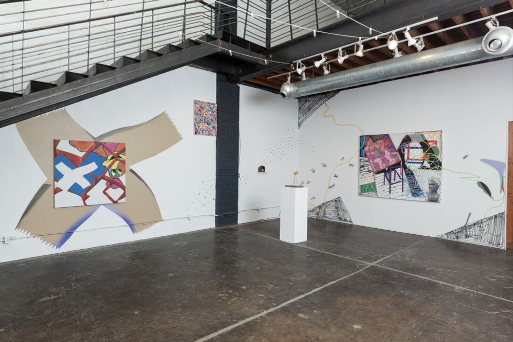 An exhibition of art in a white walled gallery with a large metal staircase. Murals are on both walls of the corner we see, paintings on top of the murals. A small sculpture sits on a pedestal and a painting is installed high on the left corner wall