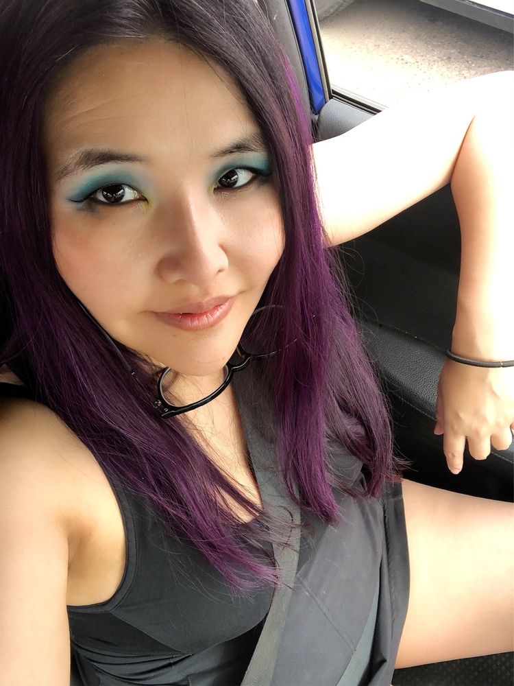 A selfie of me sitting in my car. I am wearing a black dress. I have purple hair and blue eyeshadow. 