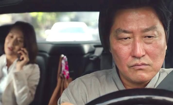 Cena de Parasita (2019) em que a mulher no banco de passageiros vê a chuva da noite anterior com positividade e o motorista, à frente, sente ódio por ter perdido tudo de sua casa com a enchente causada pela chuva