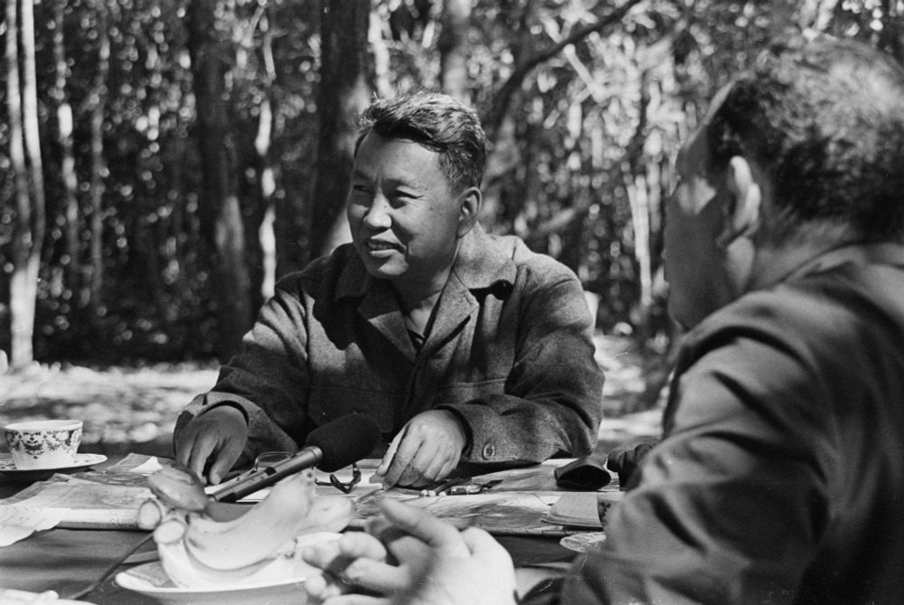Pol Pot (born Saloth Sâr), sits with other men at a table in a rural area of Cambodia, with a cup a tea.