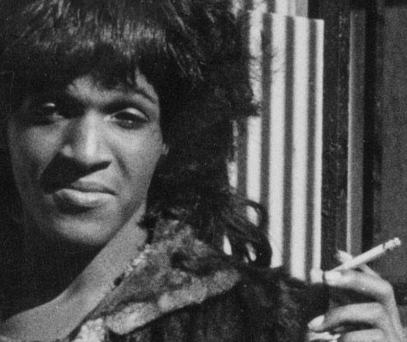 Marsha P. Johnson, wearing a fur coat, looks directly into the camera holding a cigarette between the fingers of her left hand. 