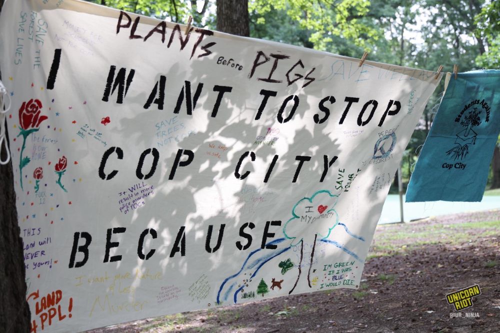 A banner hangs from a clothesline in Brownwood Park with the stenciled message “I want to stop Cop City because with many handwritten messages and drawings added on to the banner. Some messages include “plants before pigs”, “I’m green because if I was blue I would die”, “save Weelaunee” and “it WILL result in more police murders of POC”