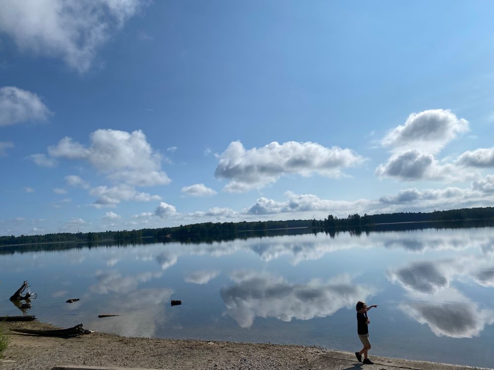 A child looks out over a calm lake in northwest Michigan. The water is as still as glass and the few clouds in the sky are reflected on the surface. 