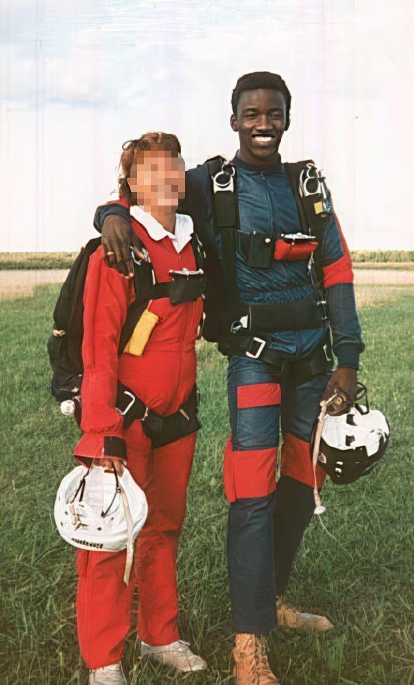 A photo of a very young Eustace (with hair!). He's a tall, slender Black man with a big smile on his face. He's standing beside another person and he's got one arm casually slung across her shoulders. They're both wearing jumpsuits--his blue, hers bright red, and they're each carrying a hockey helmet in one hand. They're both wearing parachutes--they're geared up for a skydive. I blurred her face, because I'm not sure if she'd want this like, nearly forty year old photo of herself online. But I assure you, she's smiling.
