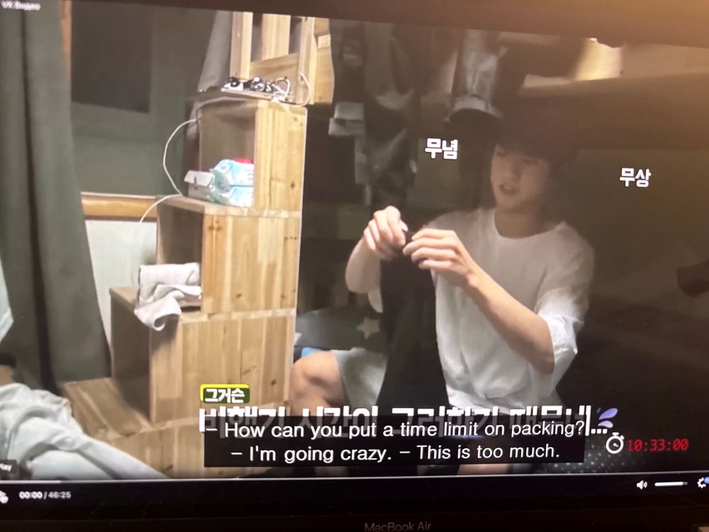 The screenshot is a frame of BTS’ Jungkook when was younger in the early days of the group’s career. He’s seen folding clothing while sitting at the bottom bunk of the dorm room, sitting beside steps that lead into the top bunk. There are subtitles that read: “How can you put a time limit on packing?”, “I’m going crazy.”, and “This is too much.”

