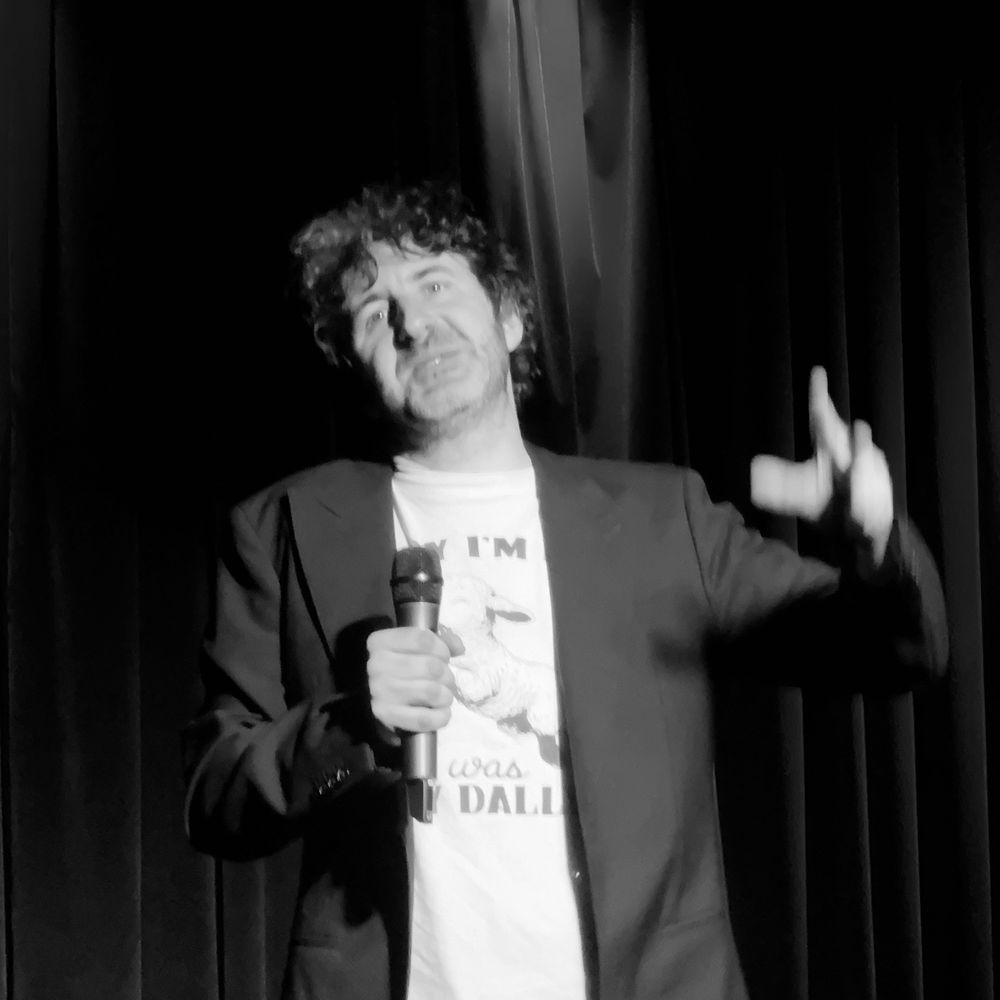 Mark Watson gesticulating on stage with a microphone, photo is monochromatic, but Mark’s emotion in his show was anything but.