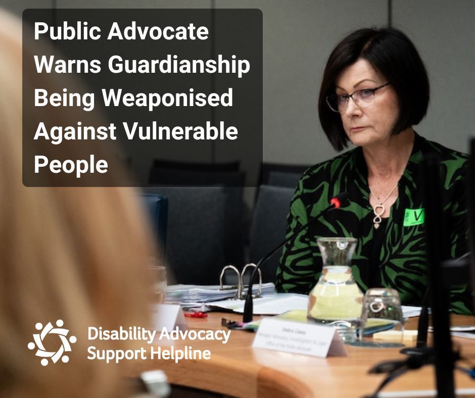 ID: A woman with short dark hair and glasses, wearing a green patterned jacket, sits at a parliamentary hearing table with documents and microphones in front of her. She appears serious and focused. A text overlay reads, “Public Advocate Warns Guardianship Being Weaponised Against Vulnerable People.”