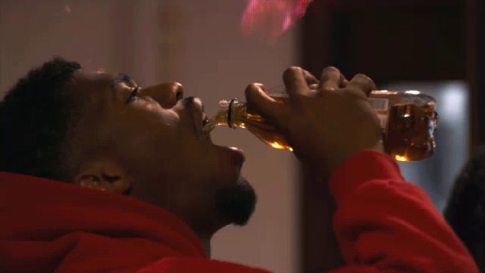 a man in a red hoodie drinks from a bottle with flames coming out of it