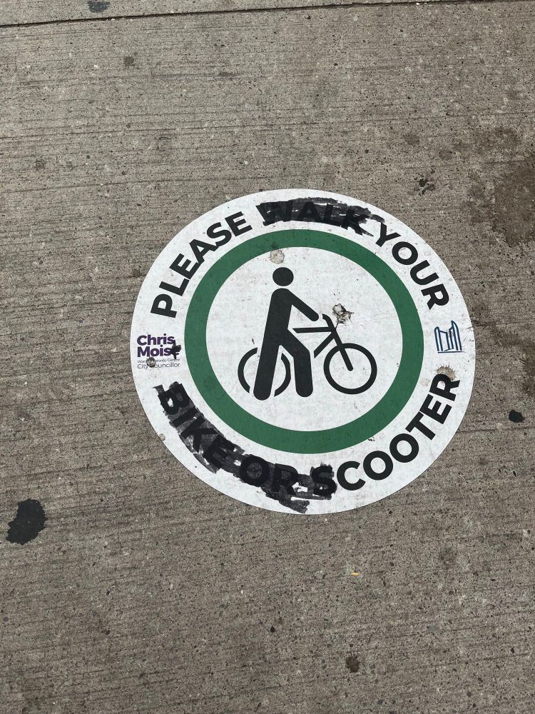 The sticker, which is round has a cyclist in the middle and was circled with the words please walk your bike or scoote where it has been “edited” to blackout some of the words and letters so now it says please your Cooter