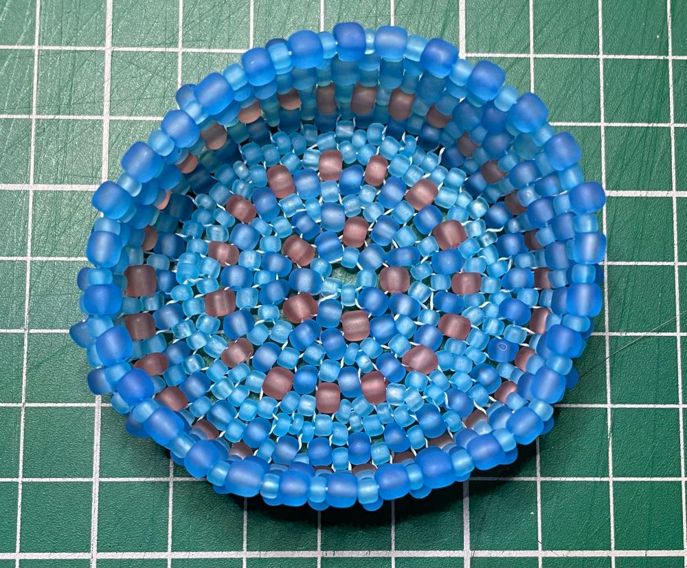 Looking down on blue and pale rose beads woven to make a shallow vessel. On a one cm green grid. 