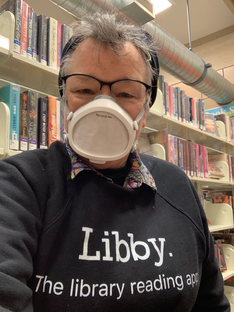 White female named Libby wearing black fleece hat, black-rimmed eyeglasses, FloMask respirator, and black sweatshirt with white lettering that reads “Libby. The library reading app.” Libby is standing in front of shelves of library books. 