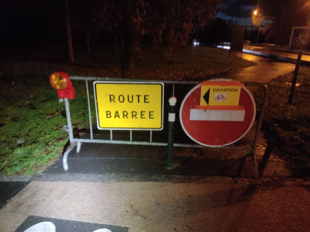 Piste cyclable fermée par une barrière sur laquelle sont apposés deux panneaux indiquant route barrée et un autre déviation 