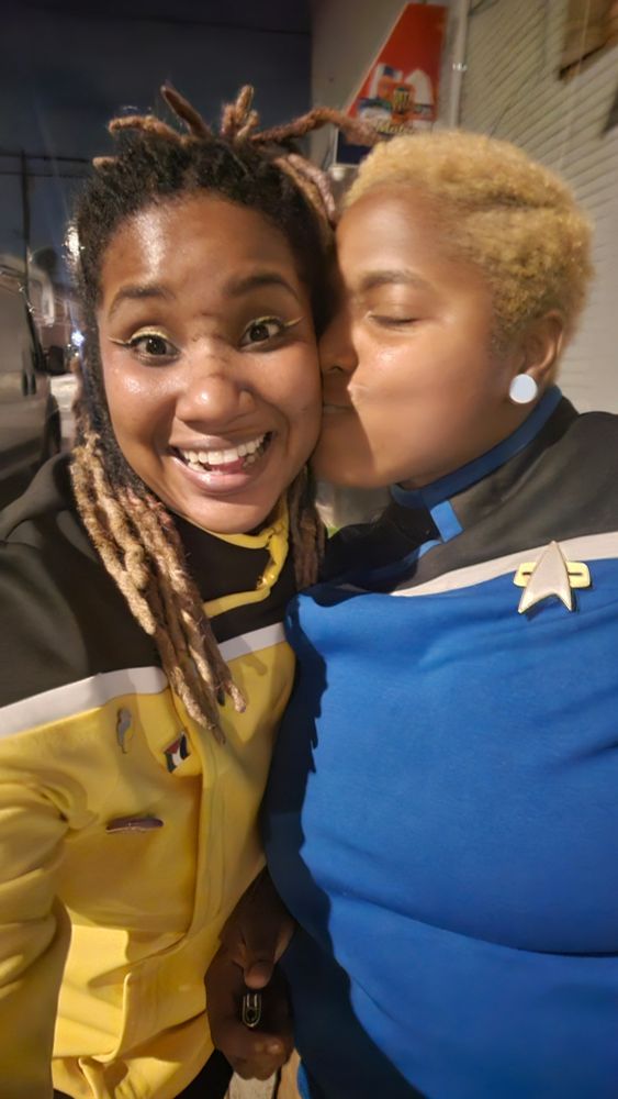 I'm smiling really big. My wife is kissing my cheek. I'm in a yellow California-class Cerritos star trek uniform. My wife is in a blue uniform of the same design with a Voyager lapel badge. 
