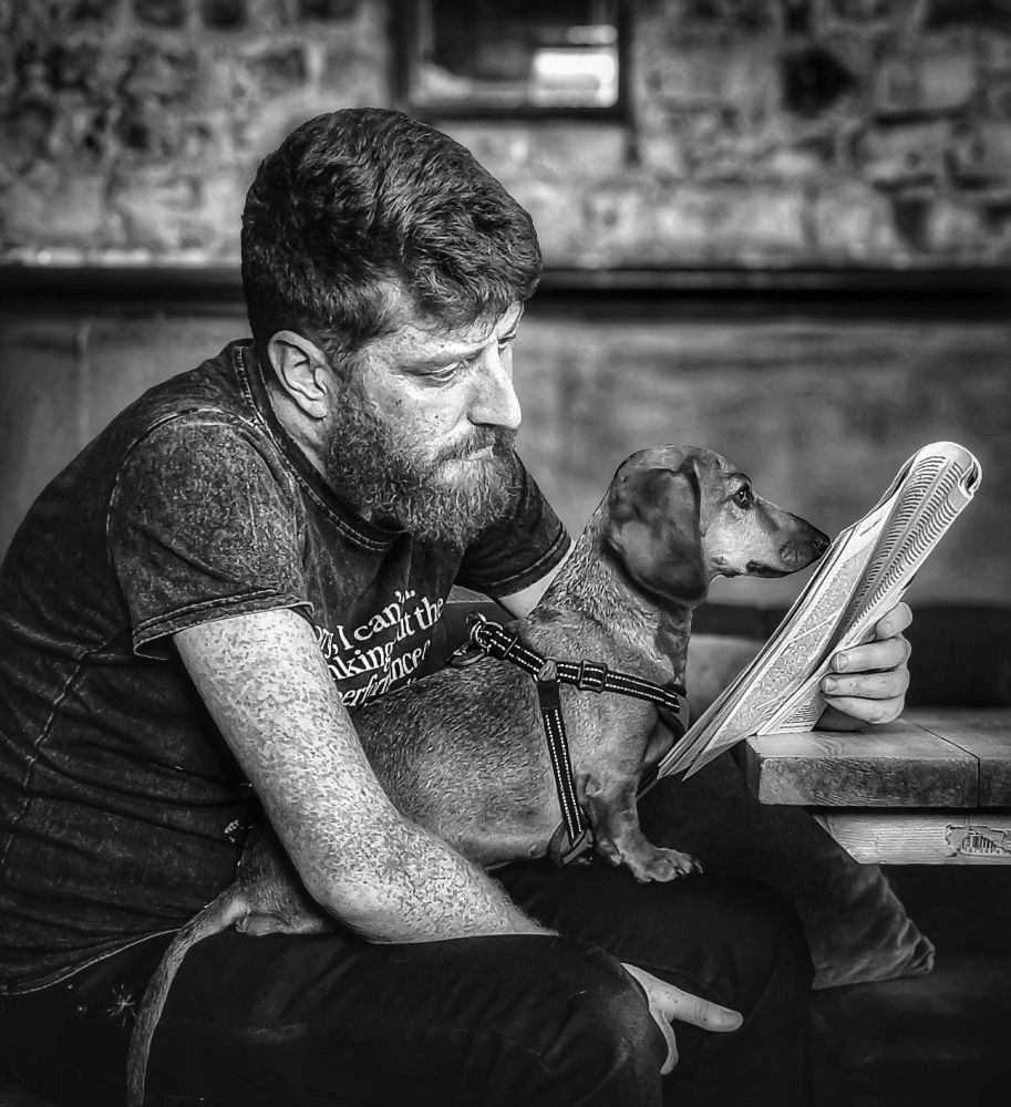 A man and his dog sit at a table in a pub and read the newspaper/journal together. 