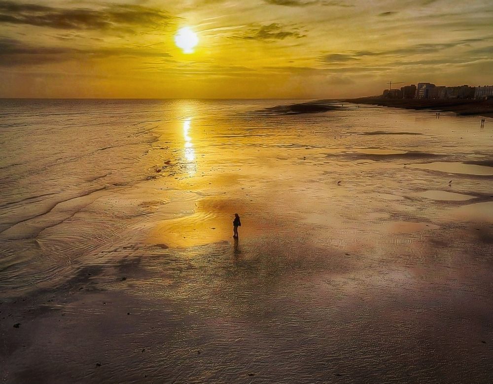 A lone figure stands on the beach looking out to sea during low tide. To the top right are buildings and then, nothing but sky and sea follows. 