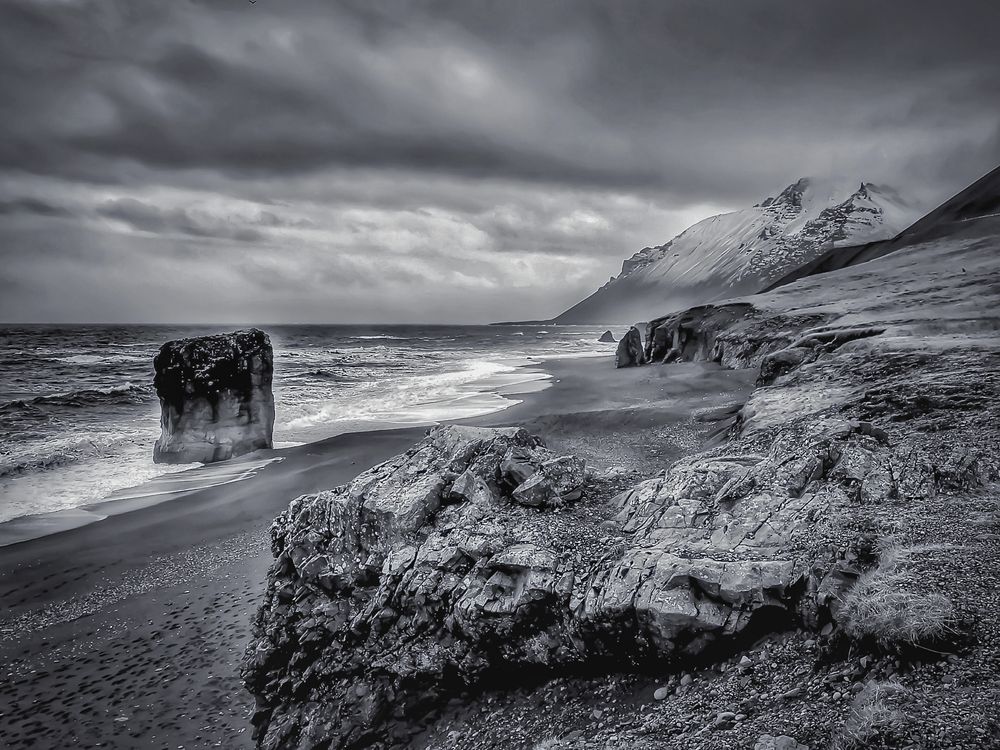 An image of a beach called Black Sand in Iceland. The shore line is rocky and in the far distance a mountain covered in snow. To the left of the image, a lone and tall rock stands jutting out from the sea. It was a stormy day and this is noticeable by the dark clouds. 
