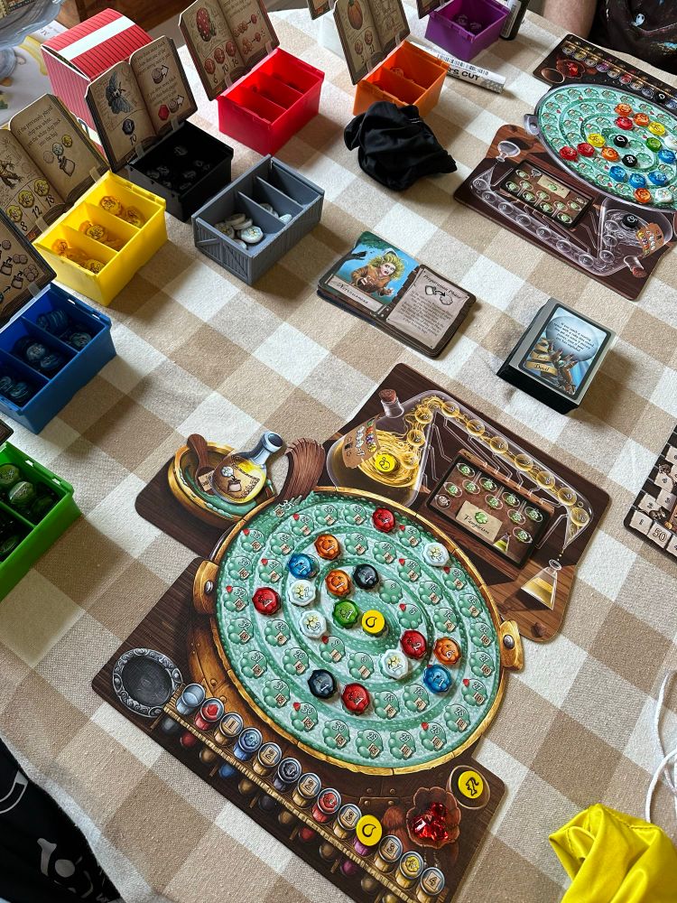A cardboard cauldron with plastic pieces on a table cloth with game cards and small plastic boxes filled with different color pieces. It’s the boardgame Quacks of Quedlinburg! 