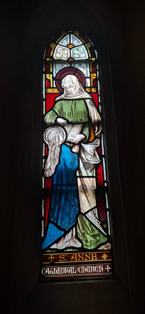 Stained glass window showing St Anne in a green dress teaching a young Mary in a blue dress how to read. 