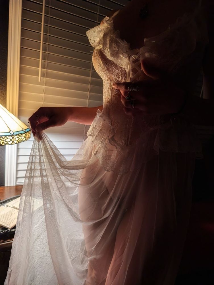 A woman in a sheer white lace dress standing in front of a lamp, the lamp illuminates her body beneath the dress, revealing her pussy and thighs 