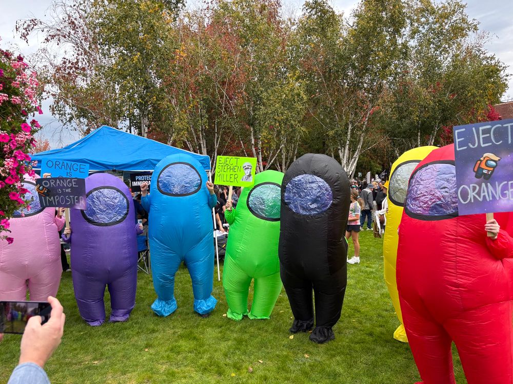 A row of AmongUs blow up costume protesters poses for a picture. There is no orange character, and the red one holds a sign that says “eject orange” and shows an orange crewmate with Trump hair and suit floating in space. 