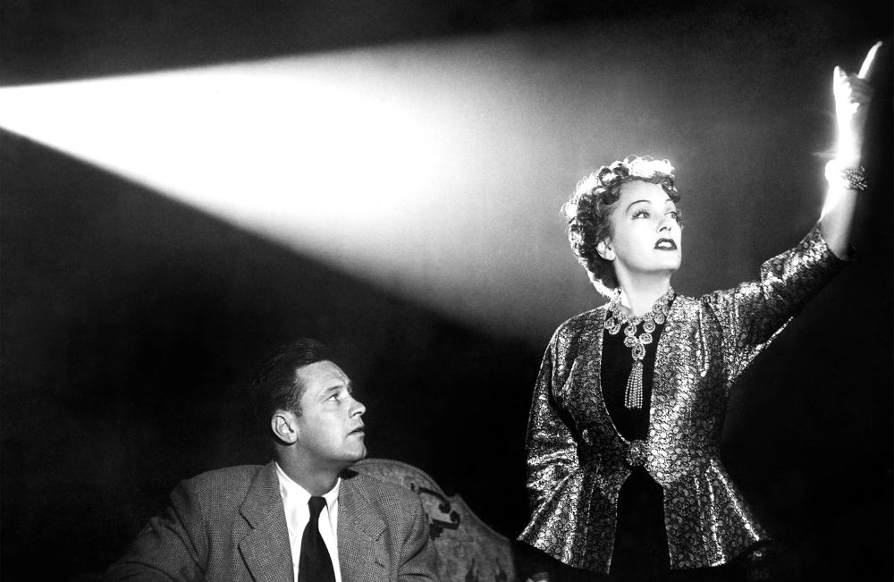 A still from Sunset Boulevard, Joe Gillis (William Holden) seated beside a standing Norma Desmond (Gloria Swanson), who holds her hand up as she stands bathed in the light of a projector