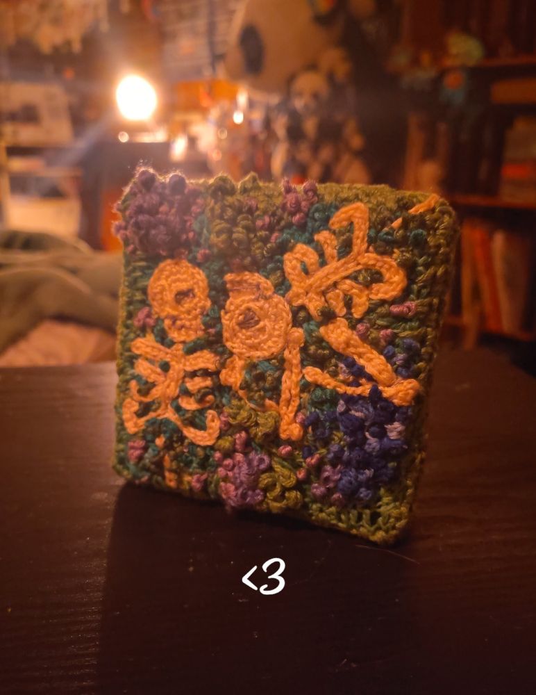 A picture of a crochet/embroidery square stretched on a canvas. The image is of 2 skeletons laying in moss and flowers, one is laying in the bottom left with its head pointed towards the top, the other is laying in the top right corner with its head pointing to the bottom left. Their heads are both in the middle, looking into each other's eyes with the bottom one's arm reaching up to hold the other's head and the other one's arm reaching up to hold the first one's arm. There's purple flowers in 2 big clumps and then also scattered around the rest of the moss and there's one big patch of blue flowers in the bottom right.