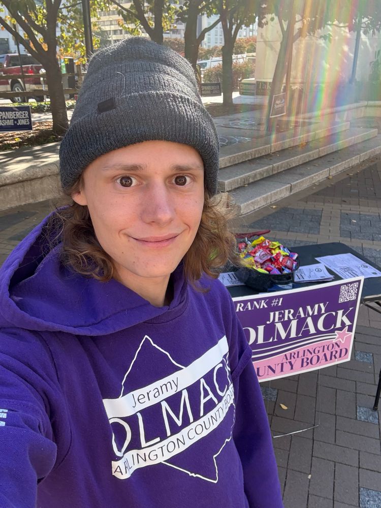 Jeramy Olmack in a grey Beanie, purple campaign shirt (with a purple hoodie underneath) standing infront of a black table with candy and campaign information.