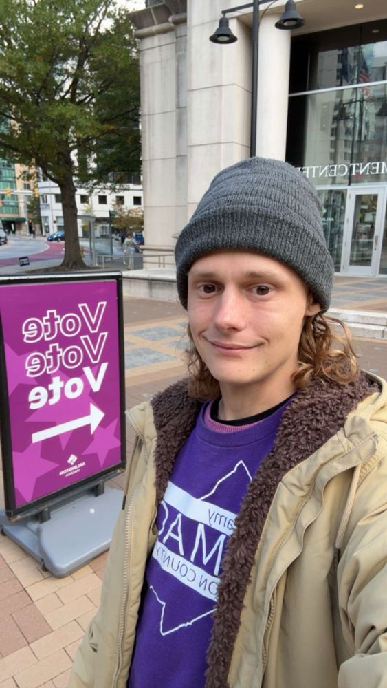 Jeramy Olmack in a beanie, tan jacket and purple campaign shirt; Outside courthouse with a purple sign saying VOTE in the background