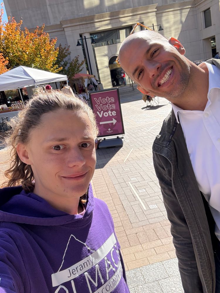 Jeramy Olmack and Carlos “DC” De Castro Pretelt standing together on Bozman Government Center Plaza with the purple vote sign in the background! 