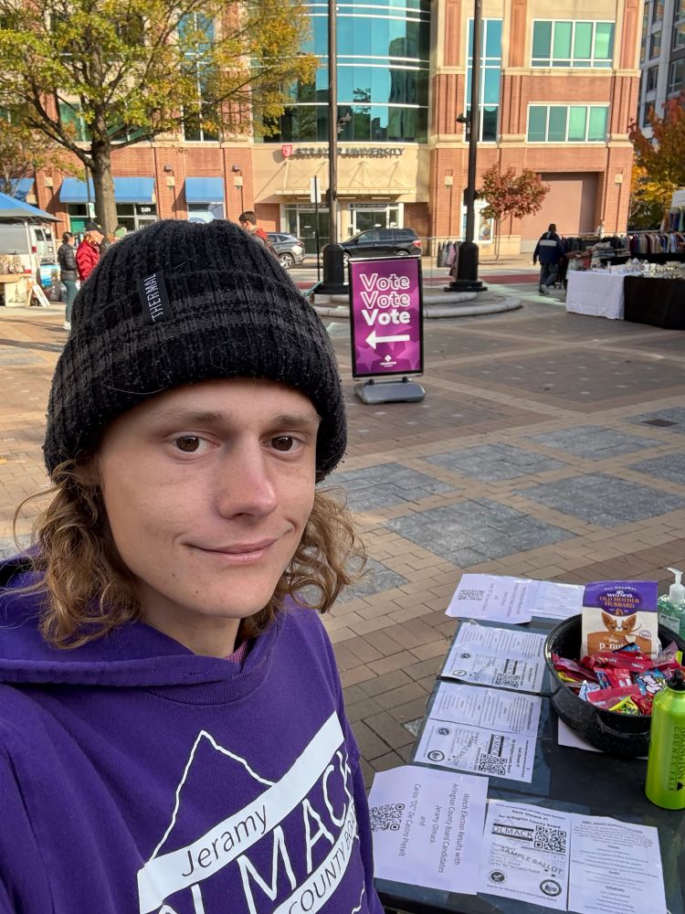 Jeramy Olmack in a black beanie, purple hoodie with a purple campaign shirt on top standing next to a black table with purple flyers and a purple vote sign in the background outside of Bozeman Government Center Plaza 