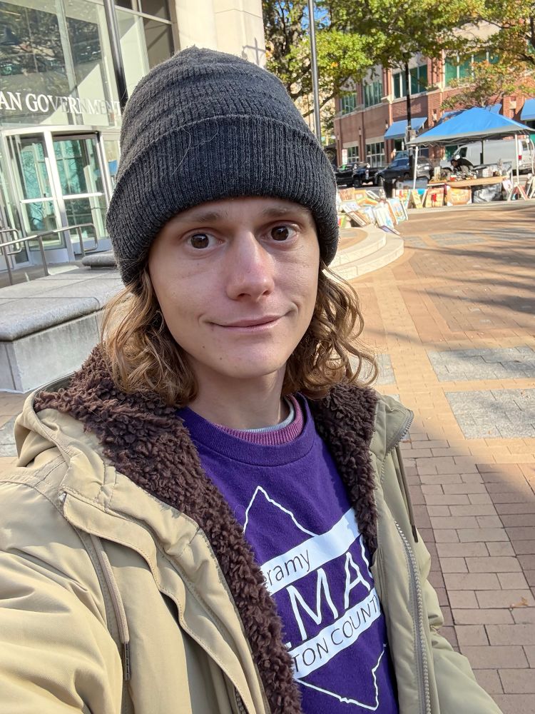 Jeramy Olmack in a tan jacket and purple campaign shirt, outside Bozman Government Center with a market in the background