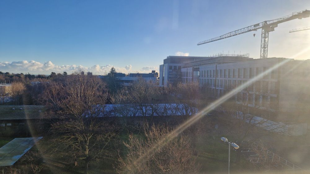 View of sunny Belfield from on top of the UCD Health Sciences Centre looking toward the North East. Bright sun in the far right corner highlights a stationary crane above the refurbishing Science building. Blue sky above gives way to low lying white clouds. In foreground are the frosty rooftops of some buildings through the tree tops.