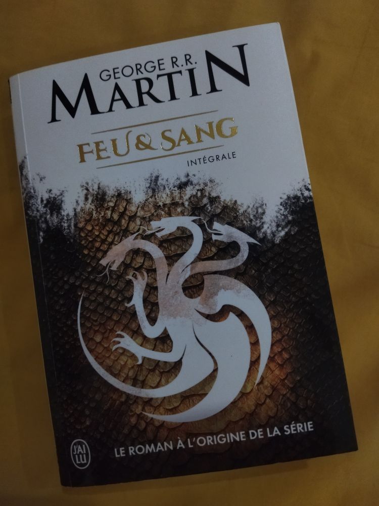 Couverture de Feu et Sang, par GRR Martin, chez J'ai Lu.
L'image est la silhouette blanche du dragon tricéphale des Targaryen sur fond d'écailles d'or et noircies. Le tiers supérieur est recouvert de blanc qui grignote l'image en direction du dragon.