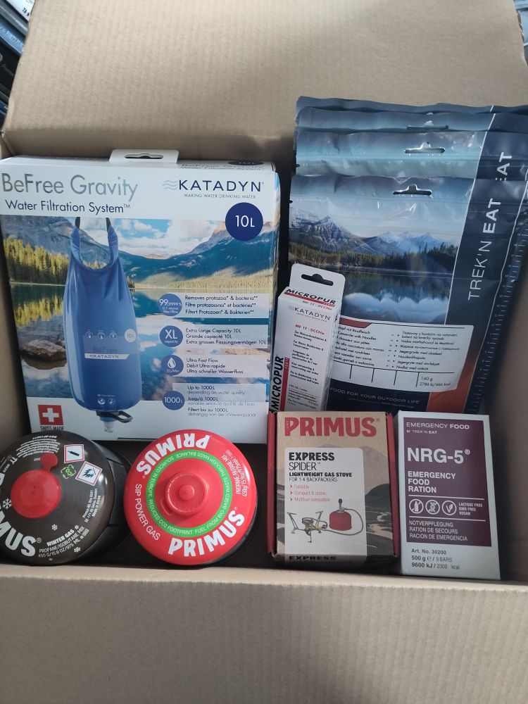 The inside of a cardboard box. It contains a water filter, a packet of water disinfection tablets, a small camping stove with two gas cartridges and some freeze-dried food packs. All items are in their original packaging as they were newly bought.