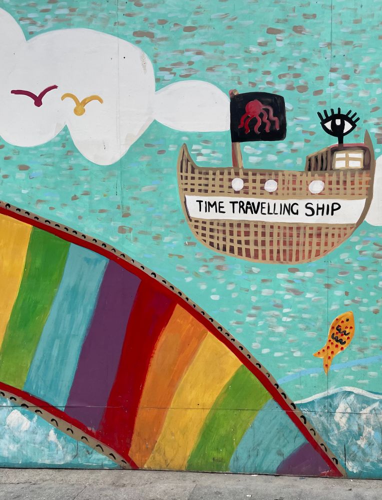 A children’s mural with a rainbow, two flying birds, a fish jumping out of the sea and a ship with a banner that says “time traveling ship”