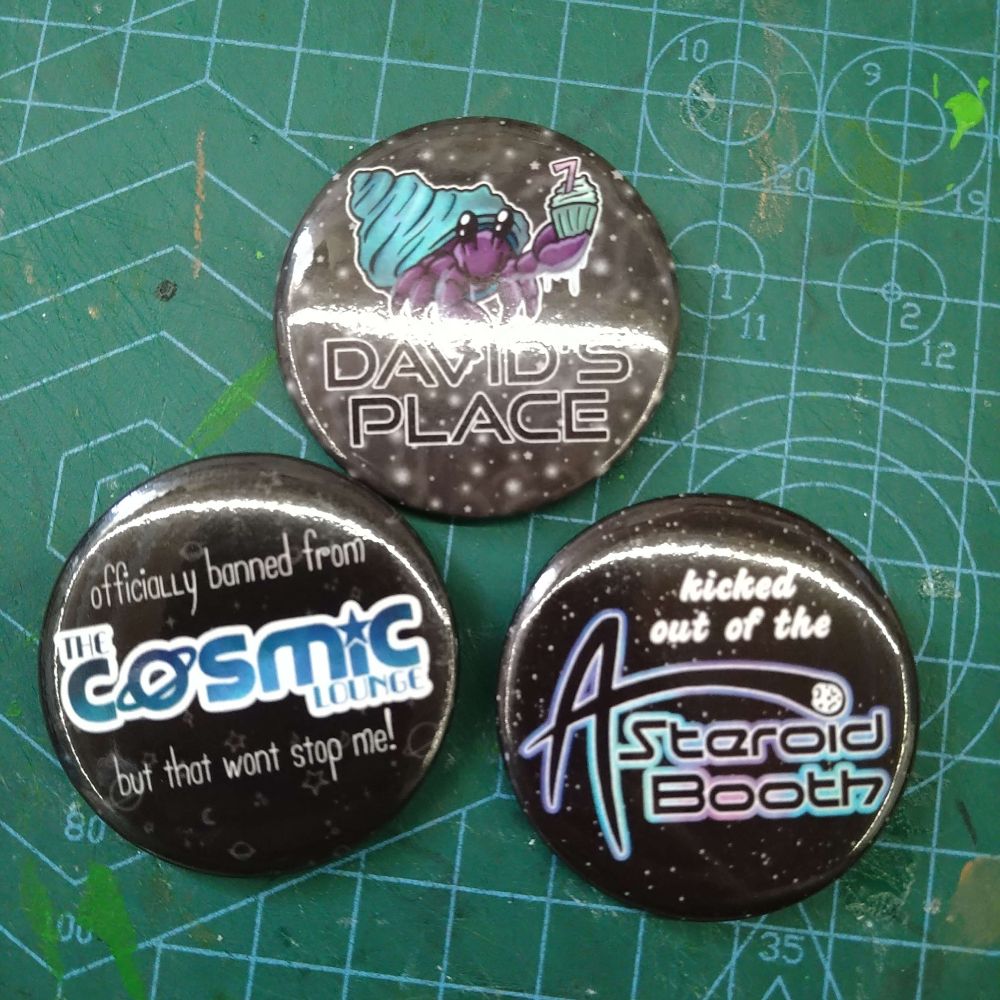 Three round badges on a green background. The first has a purple hermit crab in a blue shell holding up a cupackae with a 7 on it, and the text 'David's Place'. The second says 'Officially banned from the Cosmic Lounge but that wont stop me!'. The third says 'Kicked out of the Asteroid Booth'.