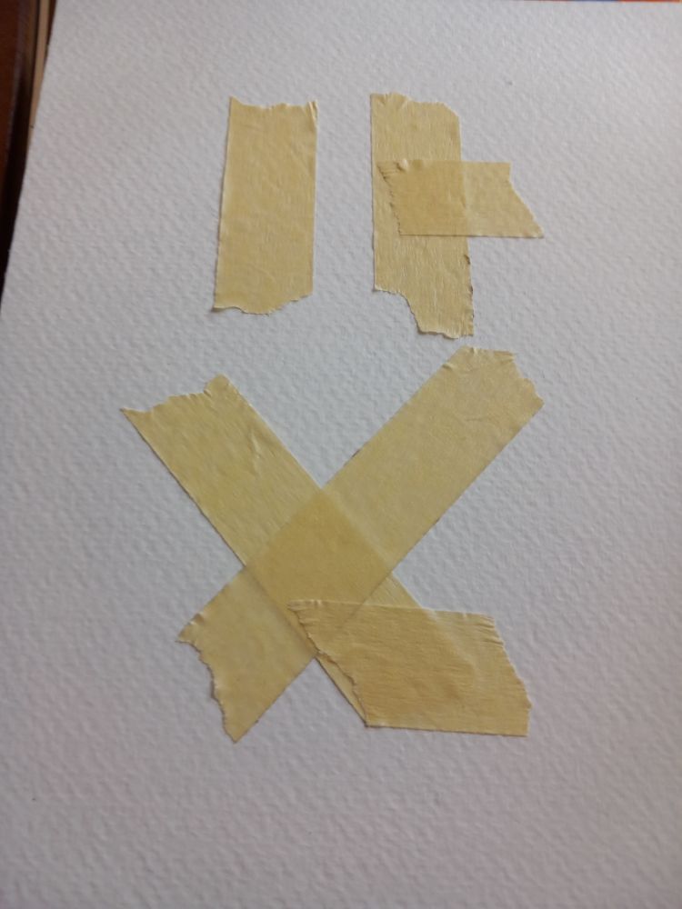 several fragments of painter's tape are arranged on a piece of watercolor paper. with a helping of pareidolia, it isn't hard to interpret the result as some sort of face displaying some sort of emotion.