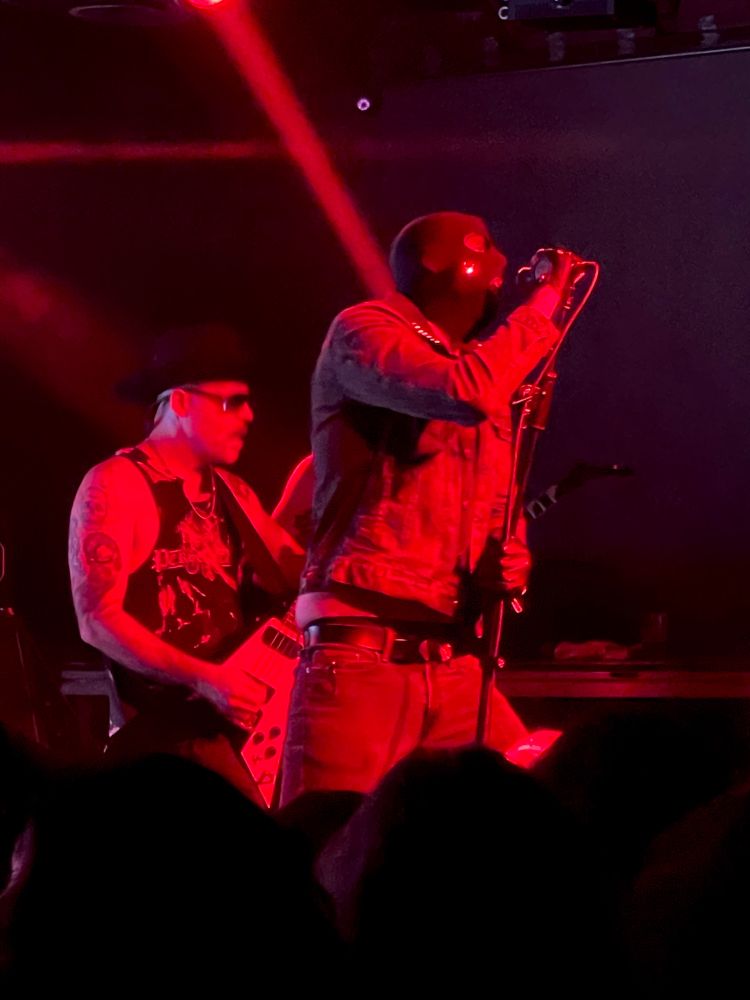 Live shot of Persekutor playing live. The bass player wears a black cowboy hat, sunglasses, and a mustache. The singer Vlad is wearing a balaclava with a bedazzled V
