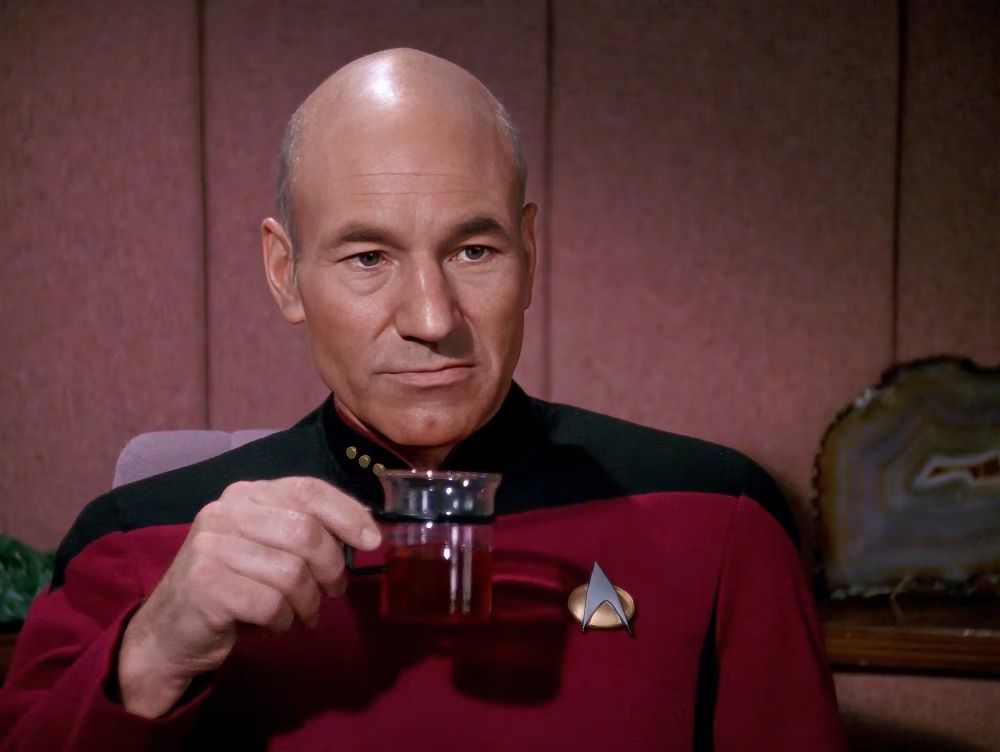 Patrick Stewart as Star Trek's Captain Picard wearing a red uniform shirt and looking at the viewer as he lifts a cup of tea