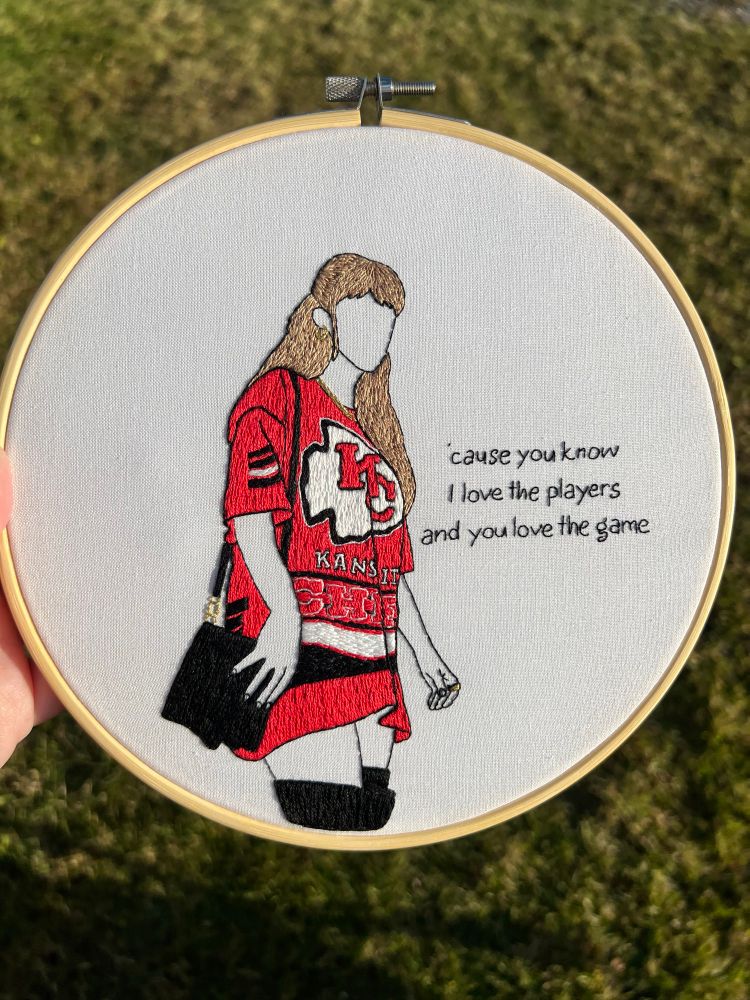 Taylor Swift hand embroidered hoop art. She’s wearing a chiefs t shirt as a dress and lyrics to blank space are next to her 
