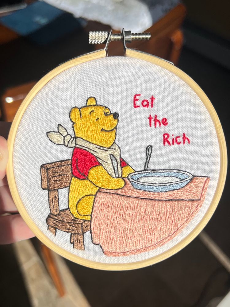 Hand embroidered hoop art of Winnie the Pooh sitting down to eat with the phrase ‘eat the rich’