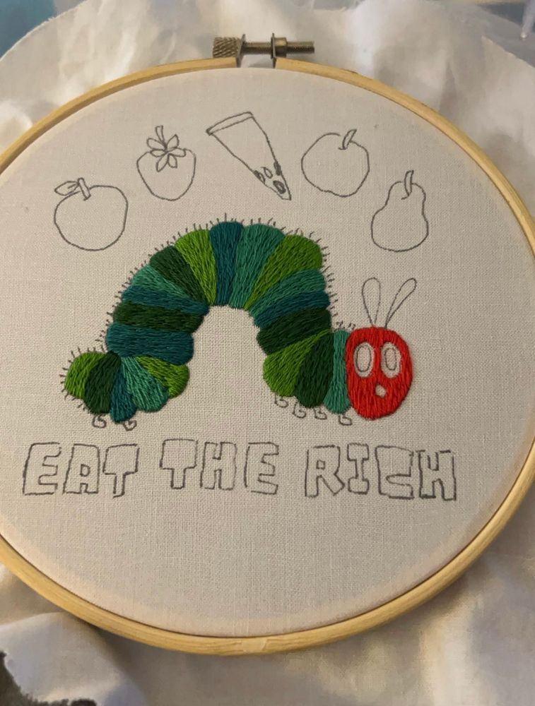 Hand embroidery in progress of the very hungry caterpillar with the phrase ‘eat the rich’ below 