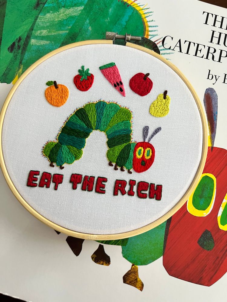Hand embroidered decor of the very hungry caterpillar with the phrase ‘eat the rich’ placed on top of the book 