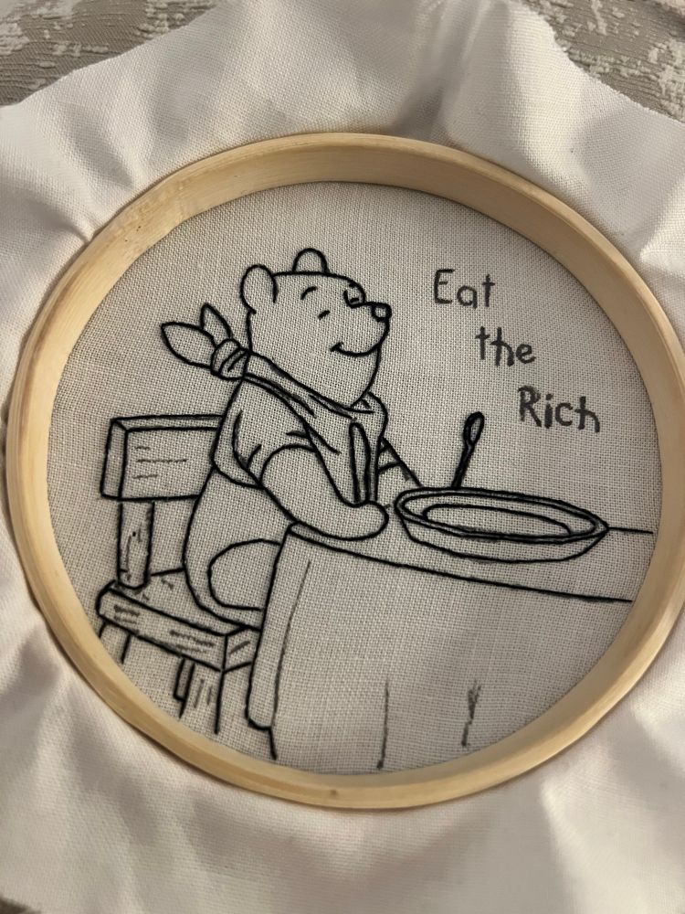 Work in progress with only the outline done, hand embroidered hoop art of Winnie the Pooh sitting down to eat with ‘eat the rich’ written on it 
