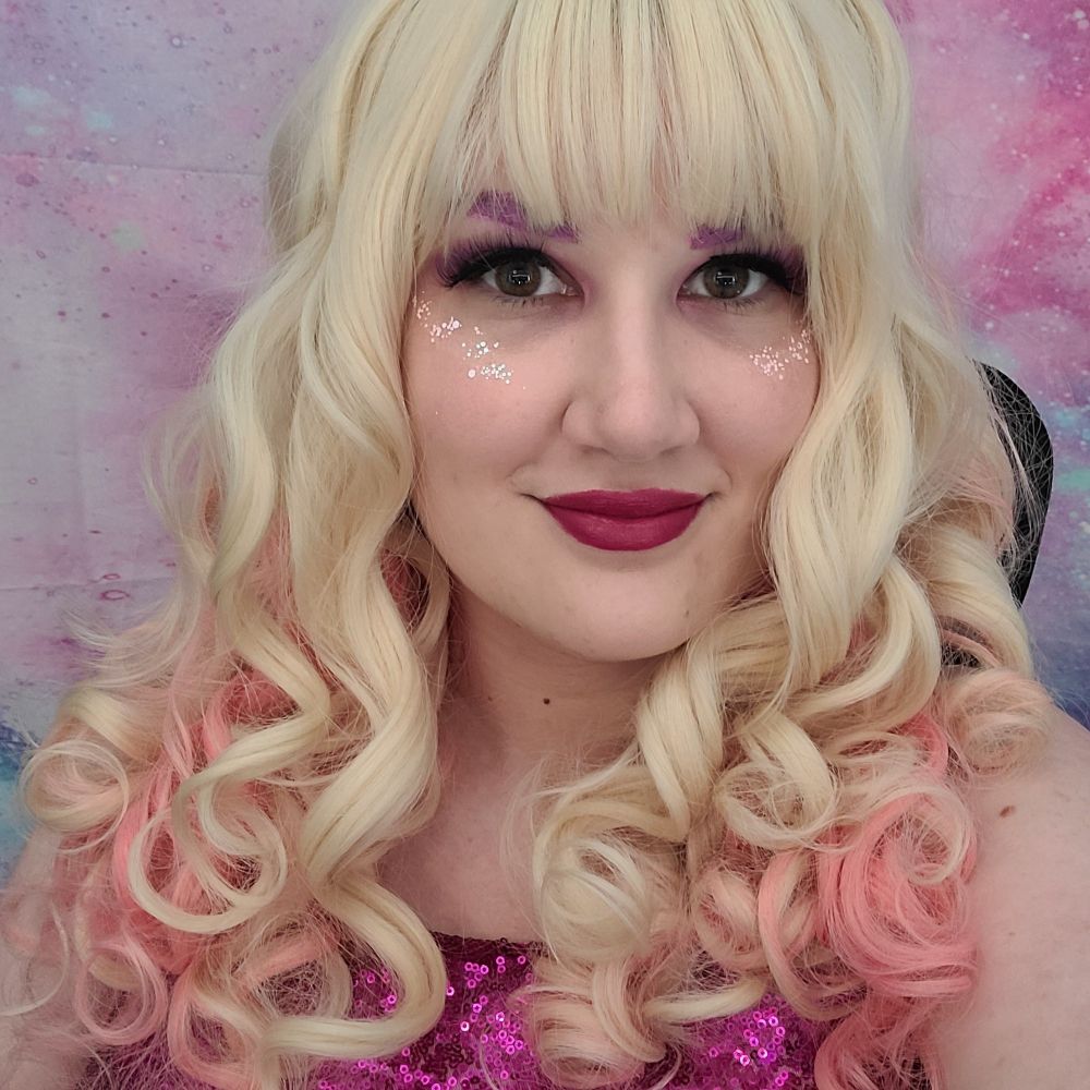 Nikki in a blond & pink wig with pink glitter on her cheekbones, pink makeup on her eyebrows, she is wearing a pink sequined top.