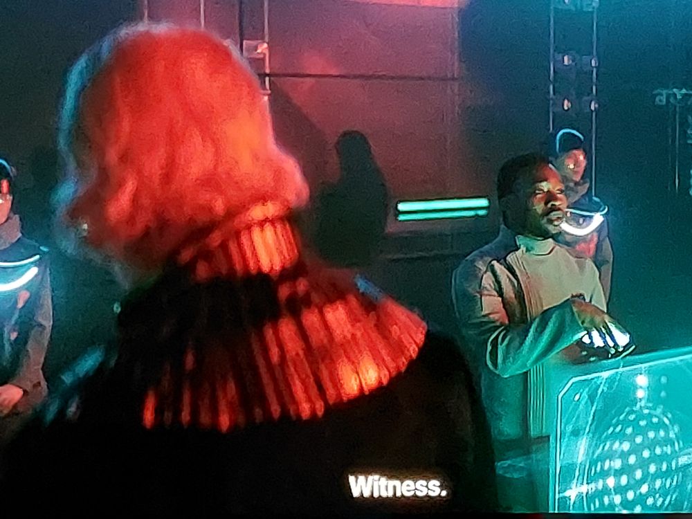 Karvis tells the character "Dawn" (Terrence Mann), "Witness" and turns a dial held in his hands. Karvis watches the screen as Dawn stands with his back to it. A green monitor holograph casts a teal light on Karvis as he stands in the background to Dawn's right. Dawn's back, facing the camera, is lit in orange by something we cannot see. The orange light glows in Dawn's white hair and glints off his gold draped shoulders.