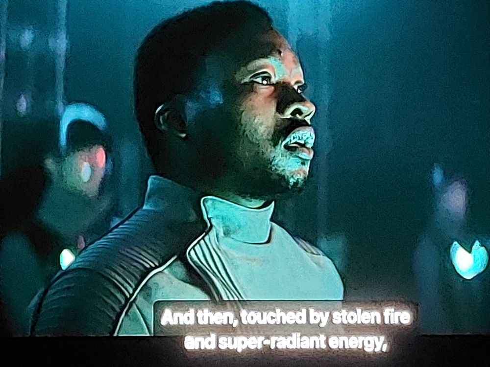 "Eely Karvis" played by Fisayo Akinade in S3E2 of "Foundation." A man answers the question "Does it work?" by describing a process of weaponry in a very poetic way. He watches the scene unfold as he describes part of it as, "And then, touched by stolen fire and super-radiant energy,". The room around him is darkly lit, two figures behind him are dimly seen. They are lit solely by something in front of them, to the image's right. They wear scifi outfits, with Karvis's a somewhat formal grey shirt with a half-colar and rippled shoulders.