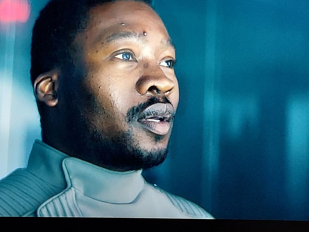 A close up of Karvis as he stares in awe at the events off screen. His face is cast in a bright white light that reflects in his eye.