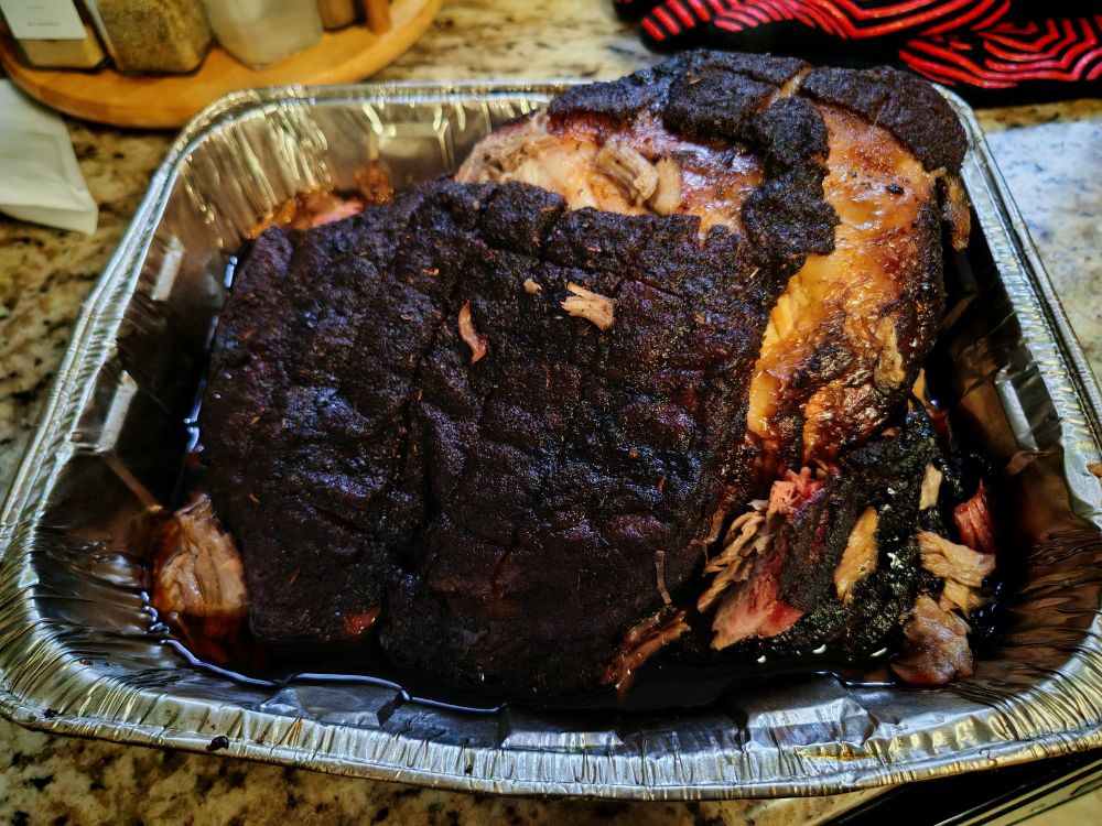 A smoked pork shoulder sitting in an aluminum tray. It is around 7 lbs and has a nice, dark bark around the outside.